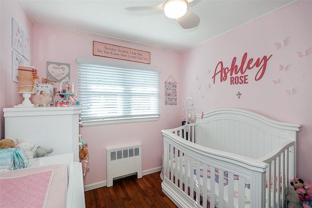 bedroom with radiator heating unit, a ceiling fan, wood finished floors, a crib, and baseboards