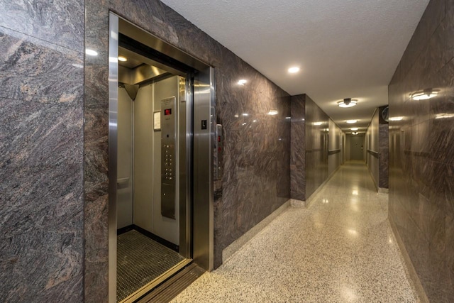 hallway with a textured ceiling, speckled floor, tile walls, and elevator