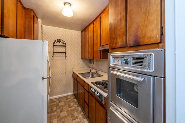 kitchen featuring wallpapered walls, appliances with stainless steel finishes, brown cabinets, under cabinet range hood, and a sink