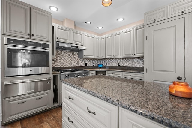 kitchen with dark wood finished floors, a warming drawer, dark stone countertops, oven, and under cabinet range hood