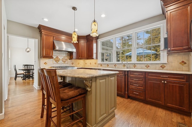 kitchen featuring a center island, decorative light fixtures, light wood-style floors, light stone countertops, and wall chimney exhaust hood