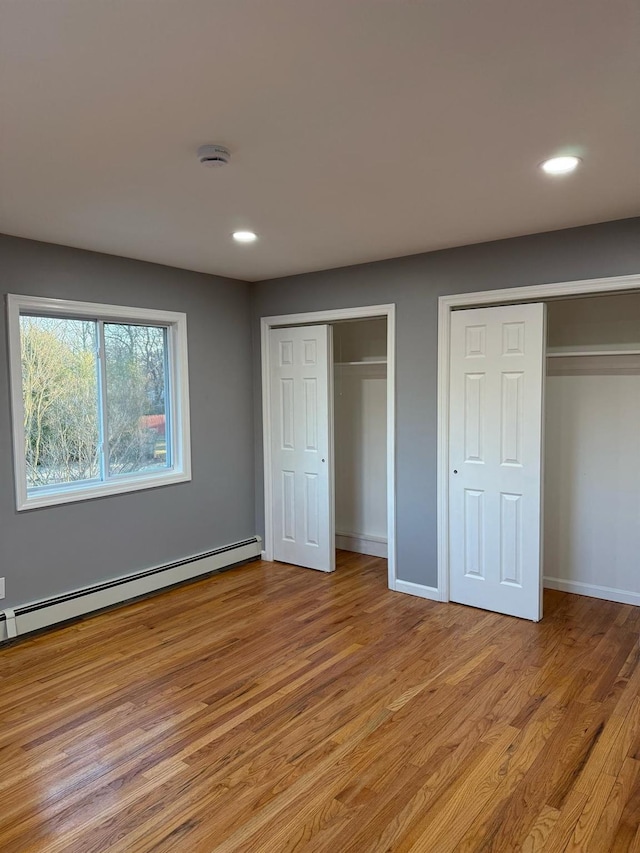 unfurnished bedroom featuring baseboard heating, light wood-style flooring, two closets, and baseboards