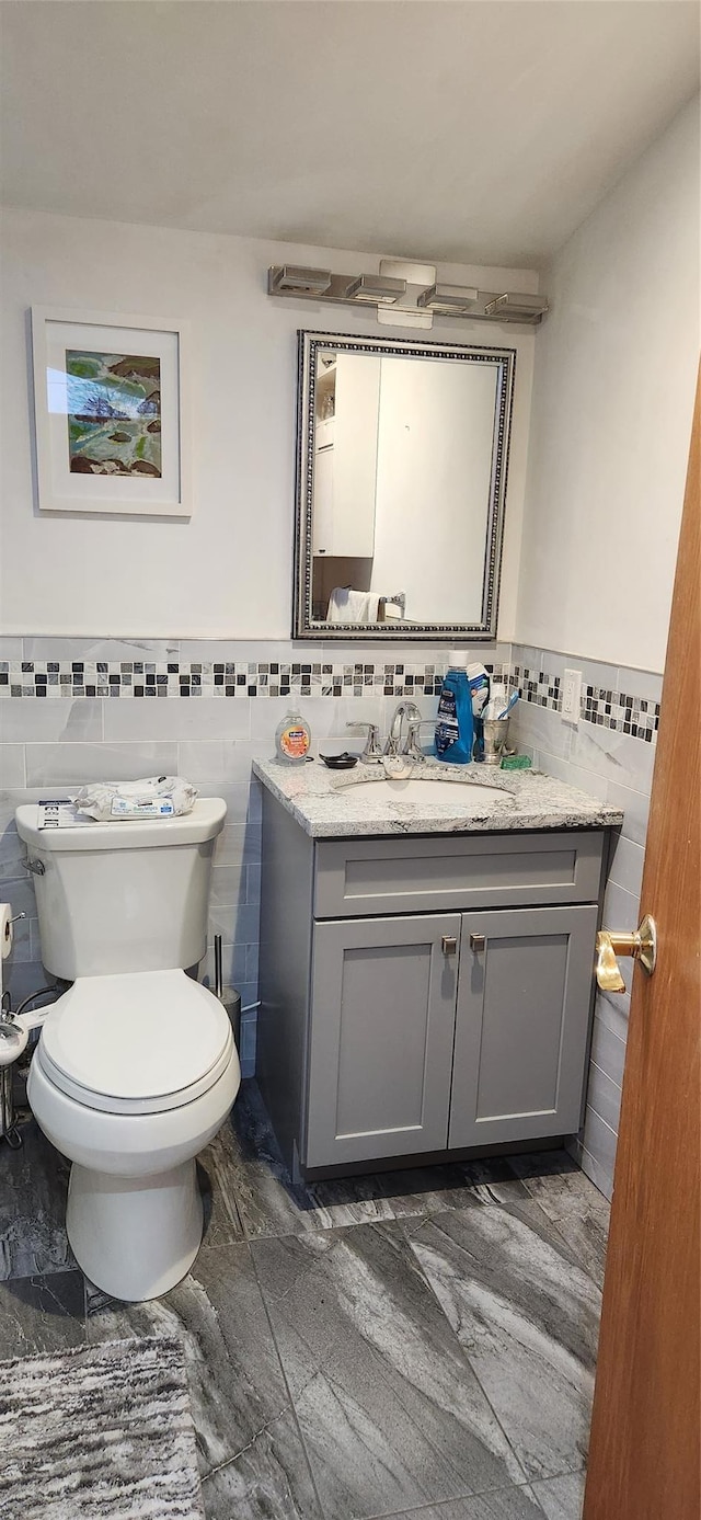 half bathroom featuring toilet, a wainscoted wall, marble finish floor, vanity, and tile walls