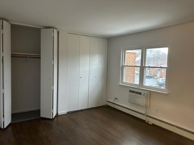 unfurnished bedroom featuring an AC wall unit, baseboard heating, dark wood finished floors, and multiple closets