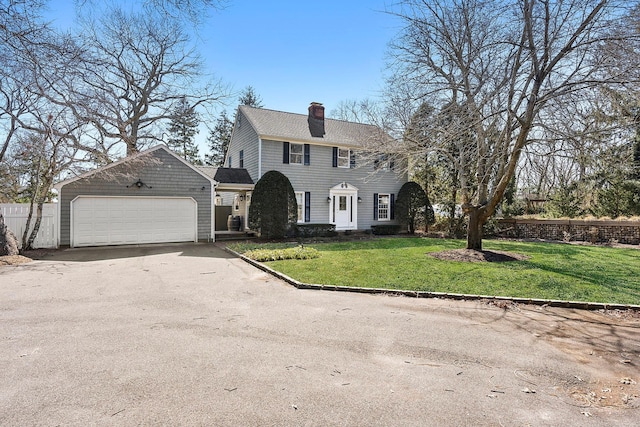 colonial-style house featuring an attached garage, a shingled roof, driveway, a chimney, and a front yard