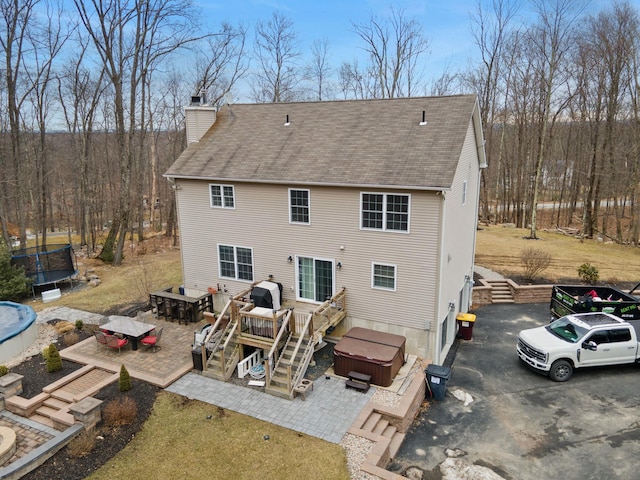 rear view of house featuring a hot tub, a trampoline, a wooden deck, roof with shingles, and a chimney
