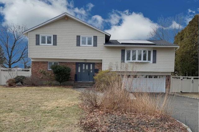 split level home featuring solar panels, aphalt driveway, fence, a front yard, and brick siding