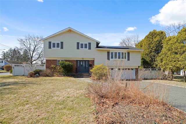 tri-level home with brick siding, fence, a front yard, roof mounted solar panels, and driveway