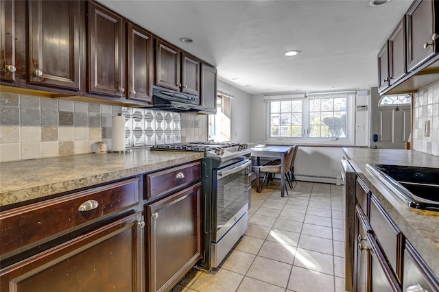 kitchen with light tile patterned floors, a baseboard radiator, under cabinet range hood, backsplash, and gas stove