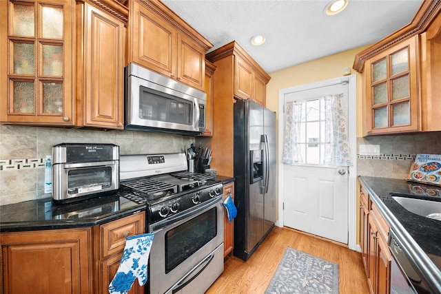 kitchen with brown cabinetry, dark stone counters, stainless steel appliances, and light wood-style floors