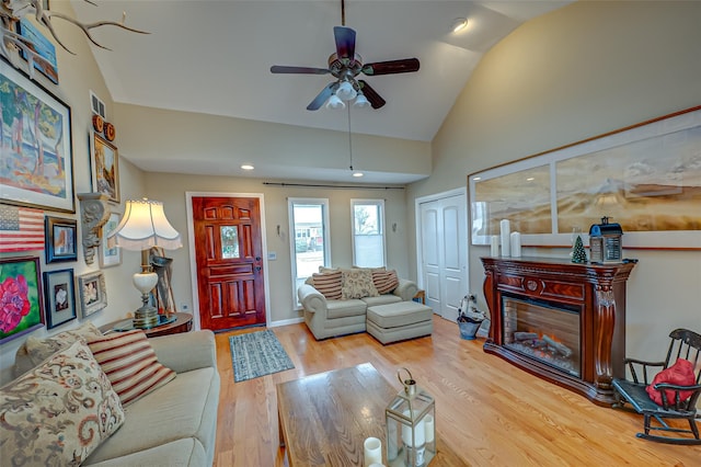 living area featuring a ceiling fan, a glass covered fireplace, wood finished floors, high vaulted ceiling, and baseboards