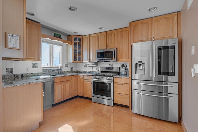 kitchen with light stone counters, a sink, appliances with stainless steel finishes, tasteful backsplash, and glass insert cabinets