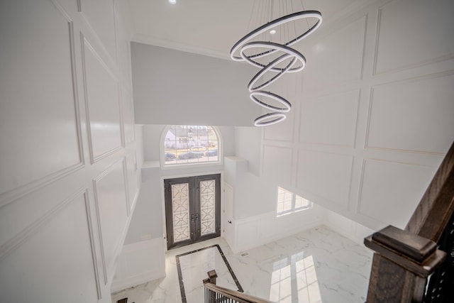 foyer entrance with a chandelier, a decorative wall, marble finish floor, french doors, and crown molding
