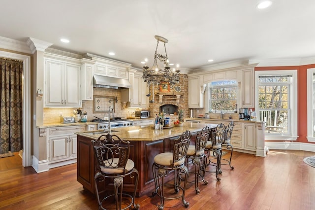 kitchen featuring a center island with sink, dark wood finished floors, light stone countertops, range hood, and crown molding