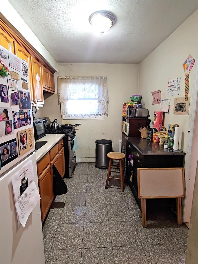 kitchen with a textured ceiling, light countertops, freestanding refrigerator, brown cabinets, and gas stove