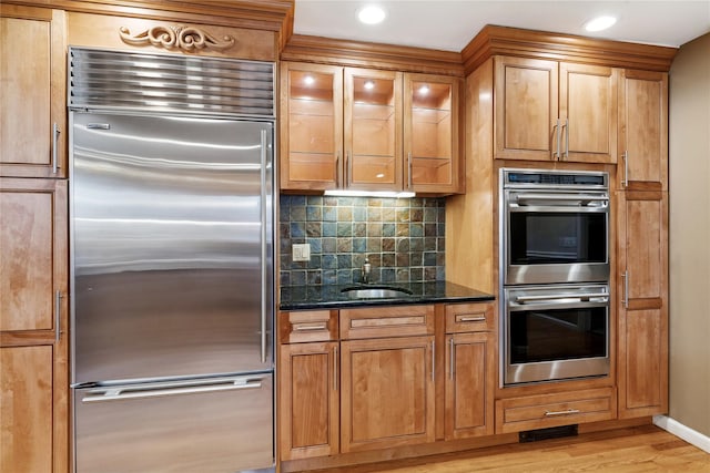 kitchen featuring light wood finished floors, tasteful backsplash, dark stone counters, appliances with stainless steel finishes, and brown cabinets