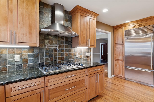 kitchen featuring arched walkways, dark stone counters, light wood-style flooring, stainless steel appliances, and wall chimney range hood