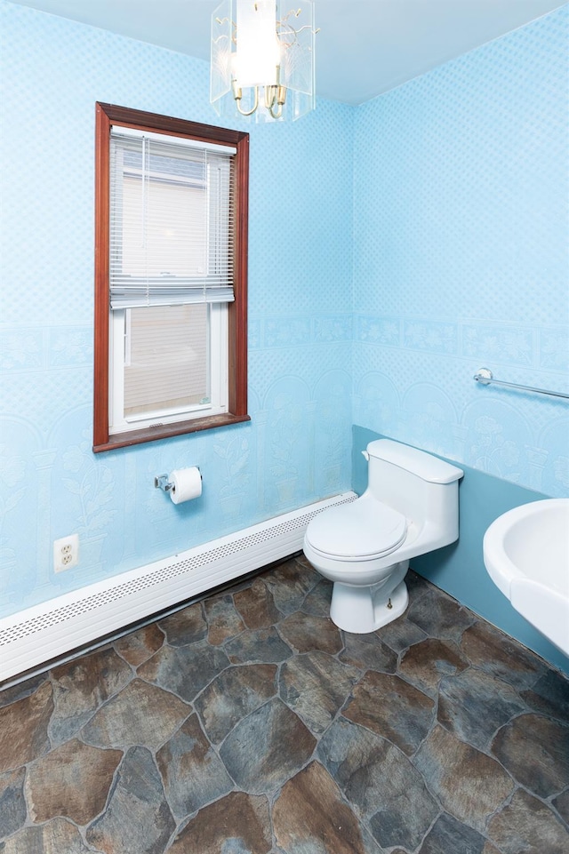 half bathroom with toilet, baseboard heating, wallpapered walls, stone finish floor, and an inviting chandelier