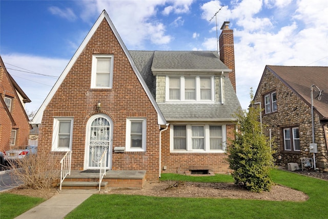 tudor-style house with brick siding, a chimney, a shingled roof, and a front lawn