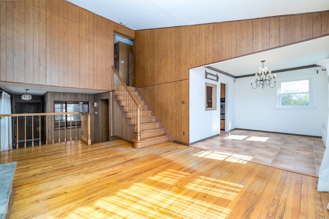 unfurnished living room featuring hardwood / wood-style flooring, wooden walls, a notable chandelier, ornamental molding, and stairway