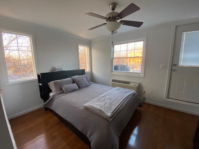 bedroom with baseboards, a wall unit AC, ceiling fan, ornamental molding, and wood finished floors