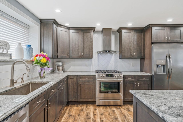 kitchen with light wood-style flooring, stainless steel appliances, a sink, dark brown cabinets, and wall chimney exhaust hood