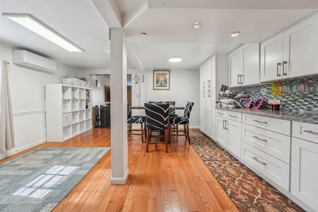 kitchen with a wainscoted wall, white cabinets, a wall mounted AC, light wood-type flooring, and backsplash