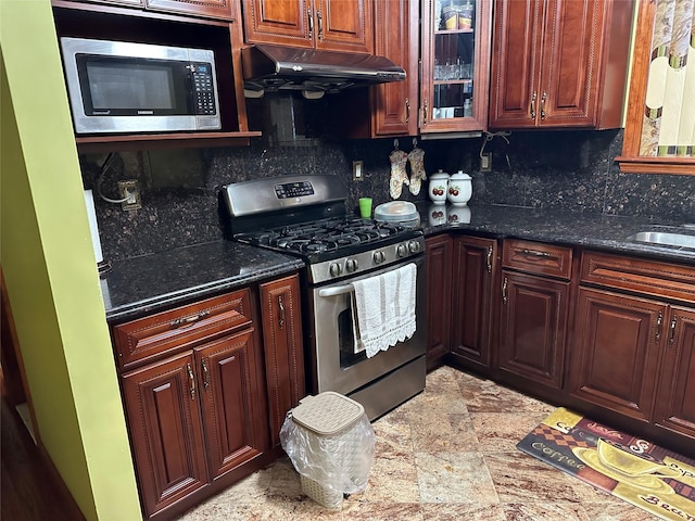 kitchen featuring under cabinet range hood, stainless steel appliances, backsplash, dark stone countertops, and glass insert cabinets