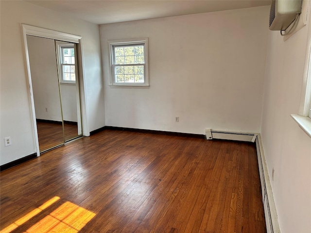 unfurnished bedroom with a wall unit AC, dark wood finished floors, a baseboard radiator, and baseboards