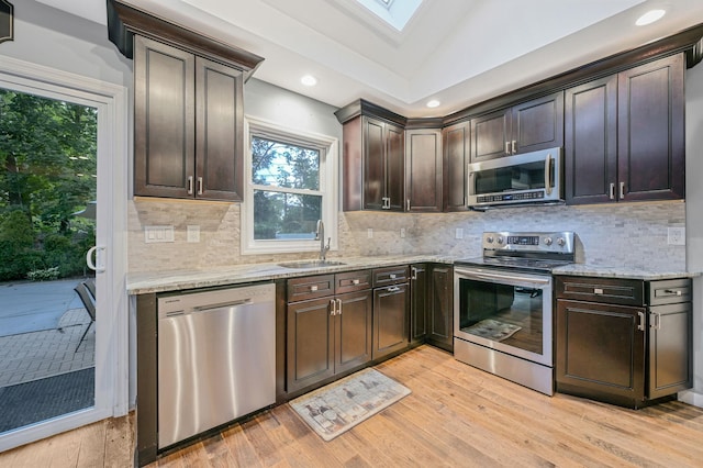 kitchen featuring a sink, appliances with stainless steel finishes, light wood-type flooring, light stone countertops, and lofted ceiling with skylight