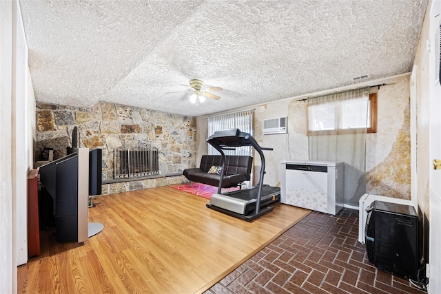 workout room with a ceiling fan, a wall unit AC, brick floor, a textured ceiling, and a stone fireplace