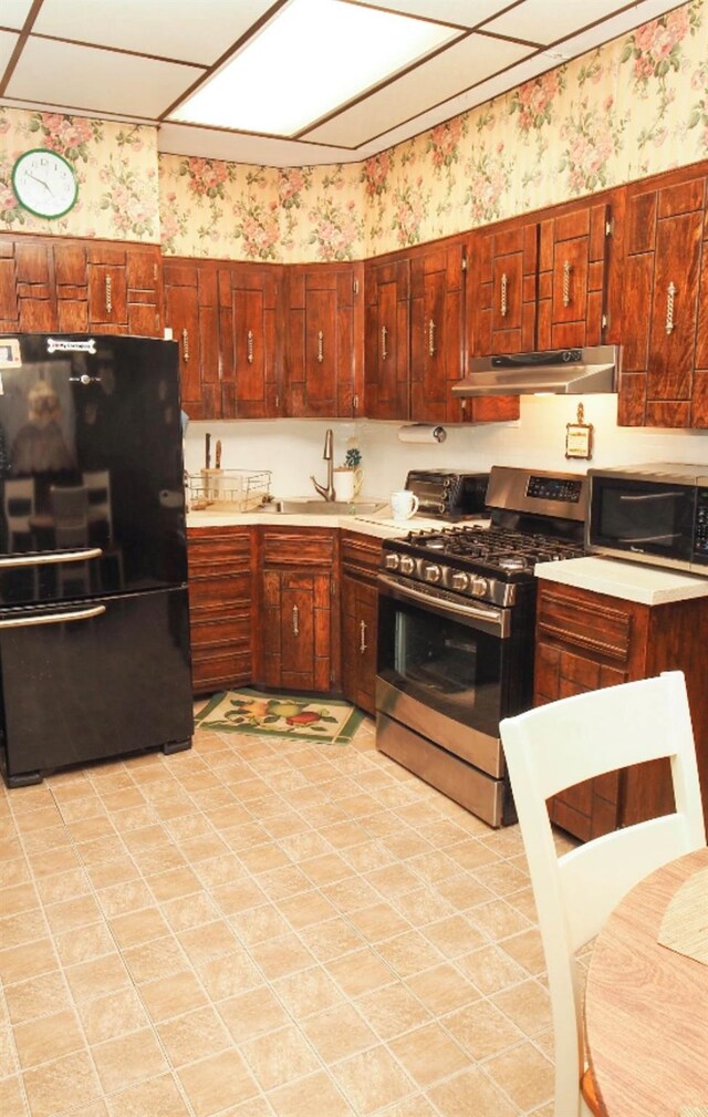 kitchen featuring freestanding refrigerator, stainless steel range with gas stovetop, a sink, under cabinet range hood, and wallpapered walls