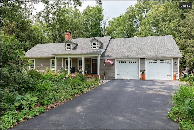 cape cod house featuring a chimney, aphalt driveway, roof with shingles, an attached garage, and a porch