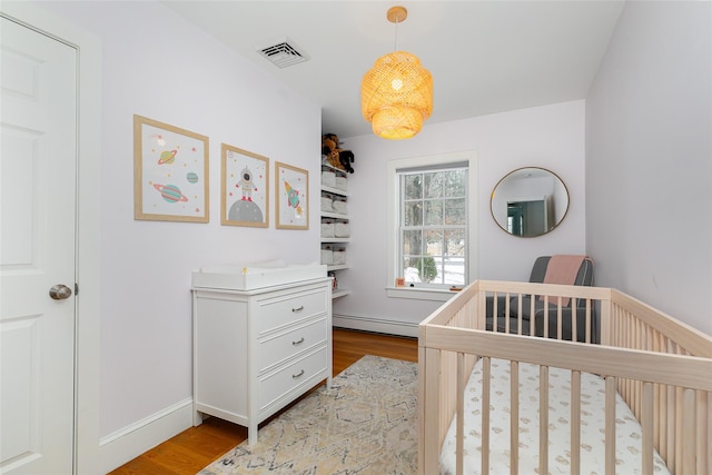 bedroom with a baseboard radiator, wood finished floors, visible vents, baseboards, and a nursery area