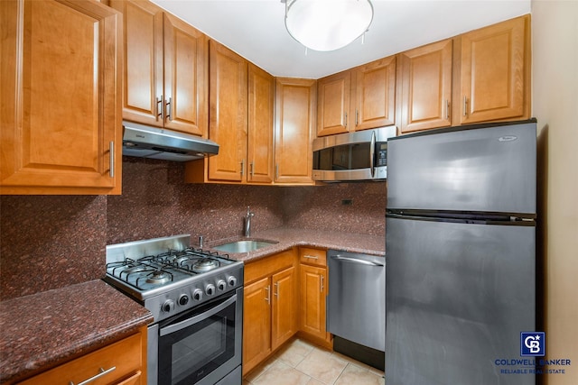 kitchen featuring under cabinet range hood, a sink, appliances with stainless steel finishes, backsplash, and brown cabinetry