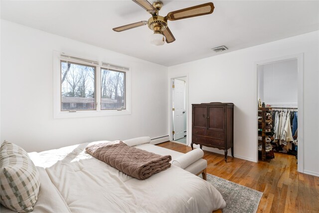 bedroom featuring a walk in closet, a baseboard radiator, wood-type flooring, visible vents, and a ceiling fan