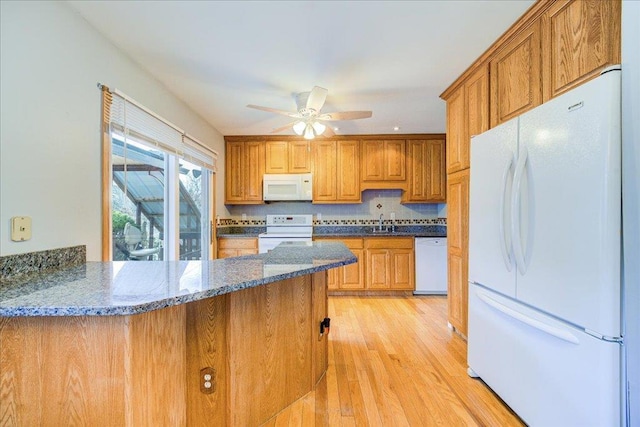 kitchen featuring dark stone counters, light wood-type flooring, brown cabinetry, white appliances, and a ceiling fan