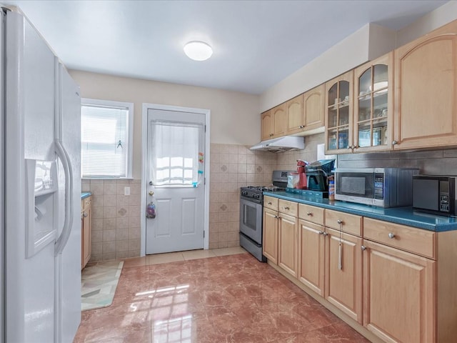 kitchen featuring tile walls, stainless steel appliances, glass insert cabinets, light brown cabinets, and under cabinet range hood