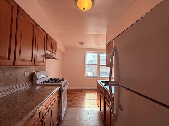 kitchen featuring brown cabinets, tasteful backsplash, white appliances, under cabinet range hood, and baseboards