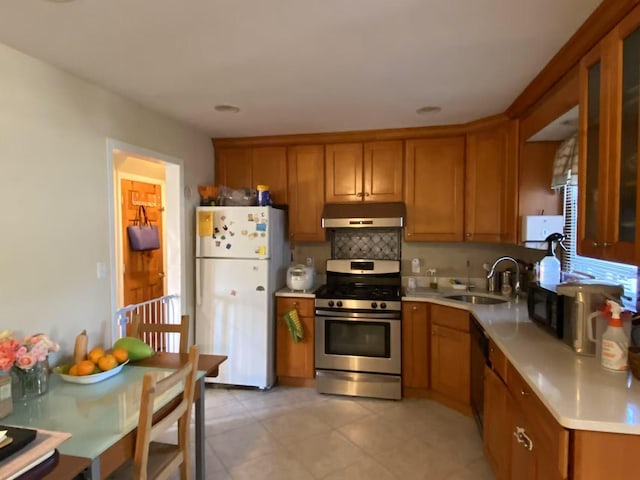 kitchen with brown cabinetry, freestanding refrigerator, stainless steel range with gas stovetop, a sink, and under cabinet range hood