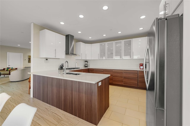 kitchen featuring light countertops, freestanding refrigerator, white cabinets, wall chimney range hood, and a peninsula