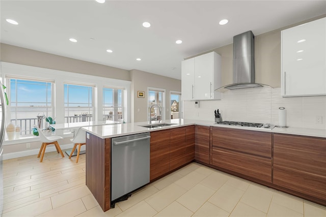 kitchen with a peninsula, a sink, wall chimney range hood, appliances with stainless steel finishes, and modern cabinets