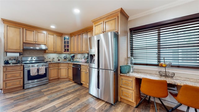 kitchen with built in desk, wood finished floors, appliances with stainless steel finishes, light stone countertops, and under cabinet range hood