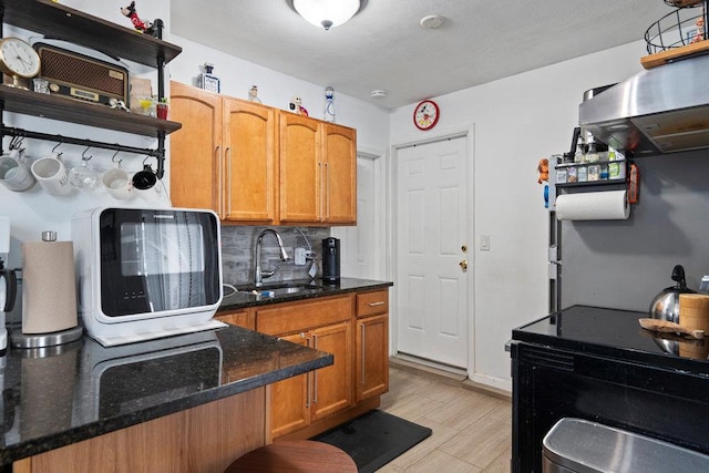 kitchen featuring light wood-style flooring, a sink, decorative backsplash, brown cabinetry, and dark stone countertops