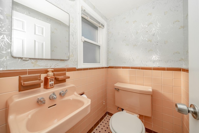 half bath with wallpapered walls, toilet, wainscoting, tile walls, and a sink