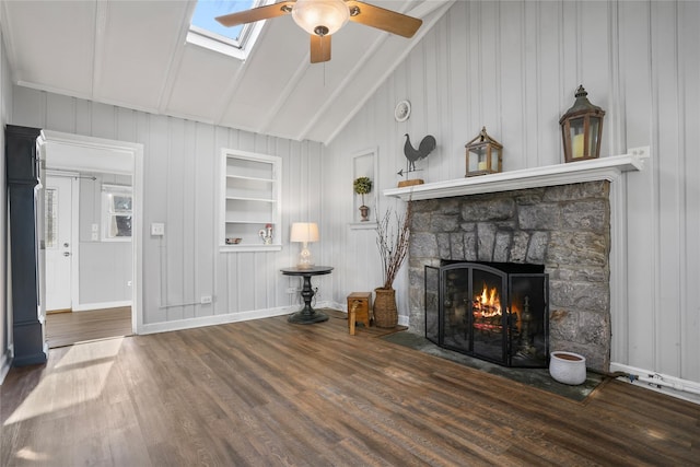 living area featuring built in features, wood finished floors, a ceiling fan, vaulted ceiling with skylight, and a stone fireplace