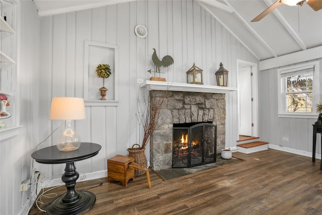 living area with lofted ceiling with beams, wood finished floors, a stone fireplace, baseboards, and ceiling fan