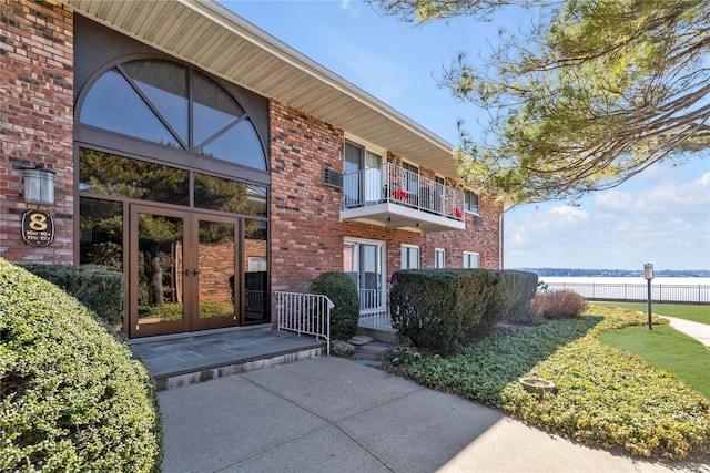 doorway to property featuring a balcony, brick siding, a water view, fence, and french doors