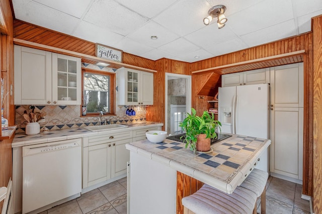 kitchen with white appliances, tile countertops, glass insert cabinets, wood walls, and a sink