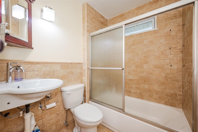 full bath featuring toilet, a wainscoted wall, bath / shower combo with glass door, a sink, and tile walls